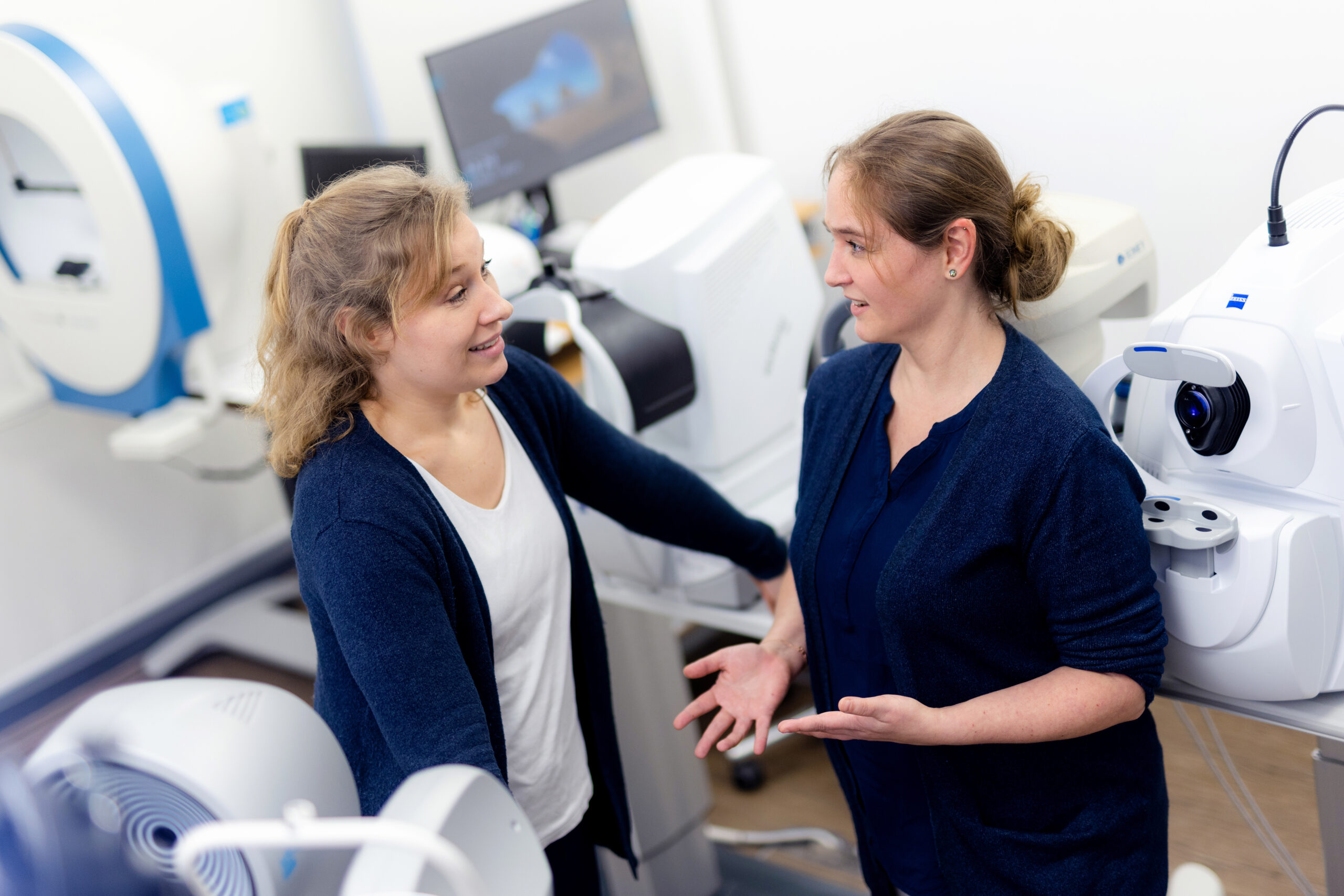 Zwei Optometristinnen unterhalten sich im Behandlungsraum zwischen verschiedenen Messgeräten.